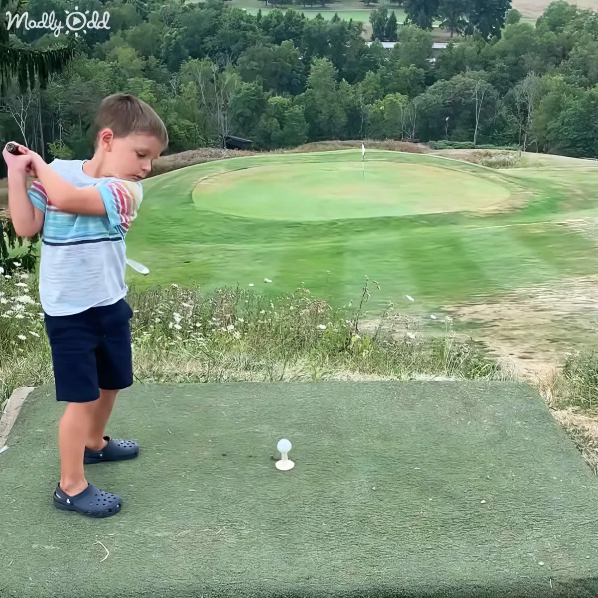 A young boy in Crocs practices his golf swing on a putting green with a distant course and trees in the background.