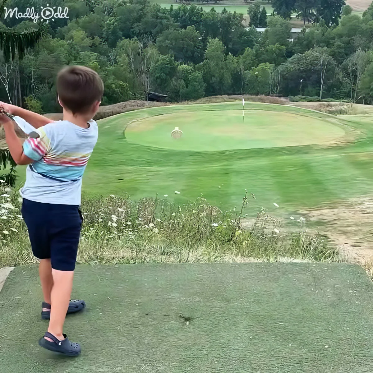 A young boy in a striped shirt swings a golf club on a driving range with a green golf course in the background.