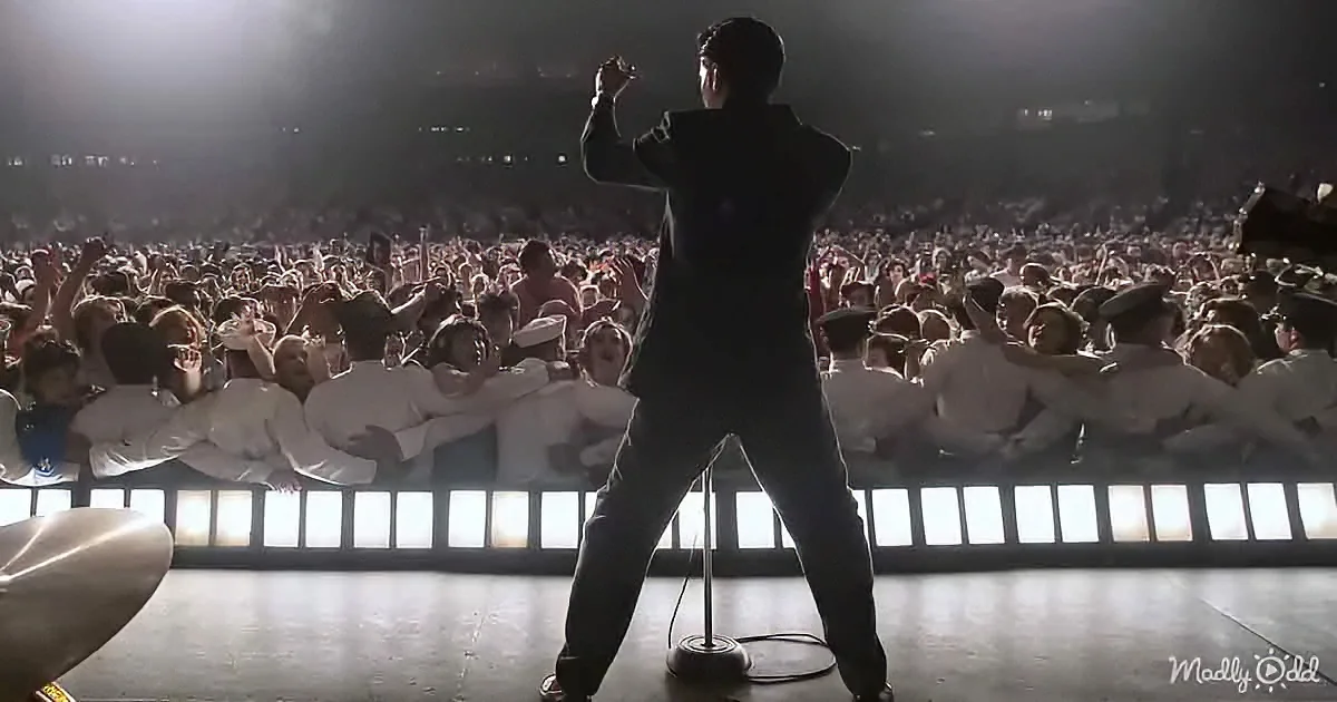 A man in a black suit stands on stage, arms raised, with a crowd of people in white uniforms behind him.