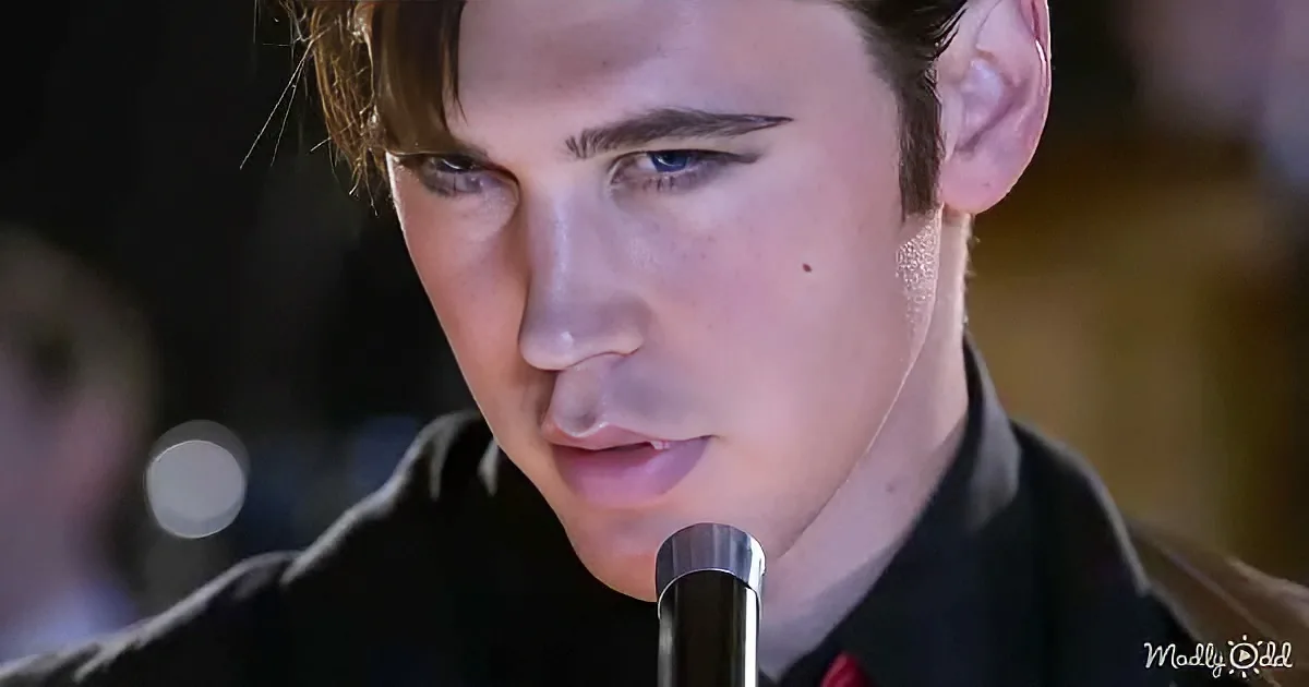 A person in a black shirt is speaking into a microphone, with a blurred background and some text in the corner.