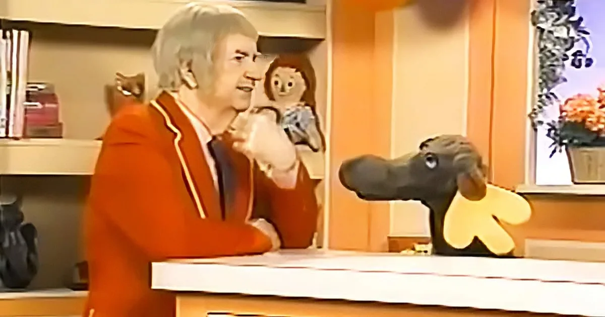 A man in an orange suit sits at a desk, gesturing with his hand near a puppet resembling a donkey.