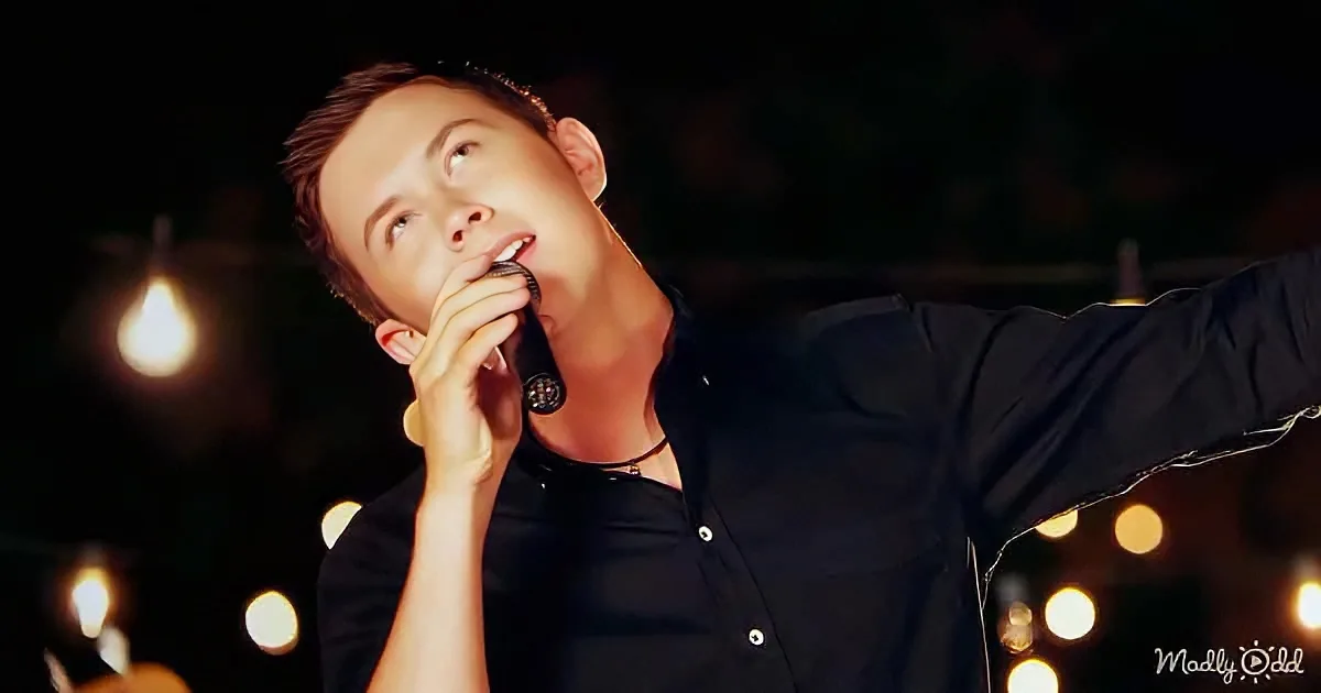A man in a black shirt sings into a microphone, arms outstretched, with blurred lights in the background.
