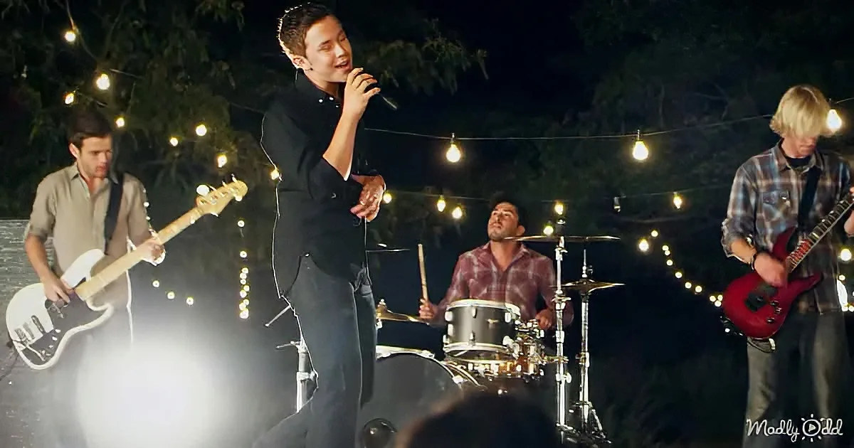 A man in black sings into a microphone while a band plays around him under string lights.