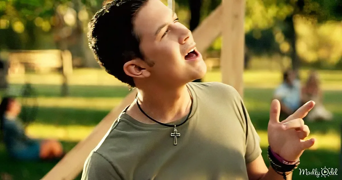 A man in a green shirt with a cross necklace, pointing upwards, in a park setting with blurred people in the background.