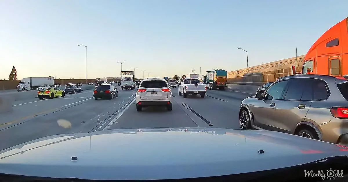 A silver car drives on a busy highway with various vehicles, including a yellow sports car and a white truck, under a...