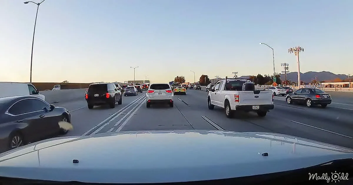 A white car drives on a busy highway with multiple lanes of traffic, under a clear sky.