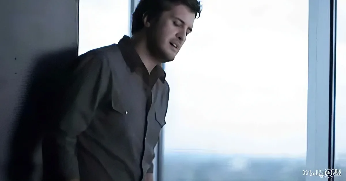 A man in a dark shirt leans against a wall, looking out a window with a contemplative expression.