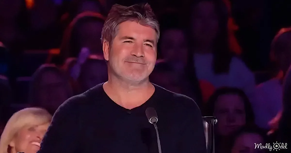 A man in a black shirt smiles while seated in front of a microphone, with a blurred audience in the background.