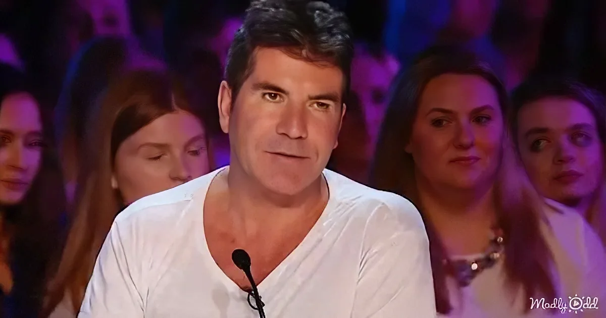 Simon Cowell in a white shirt, seated, with a microphone, in front of a blurred audience.