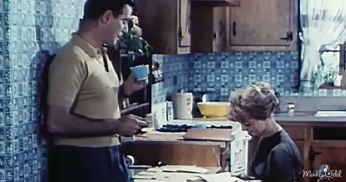 A man in a yellow shirt stands in a kitchen, holding a cup, while a young boy sits at a table, focused on something.
