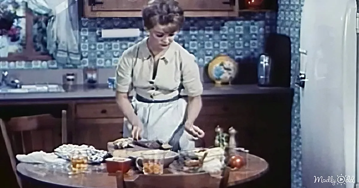 A woman in a kitchen wearing an apron, preparing food at a table with various items.