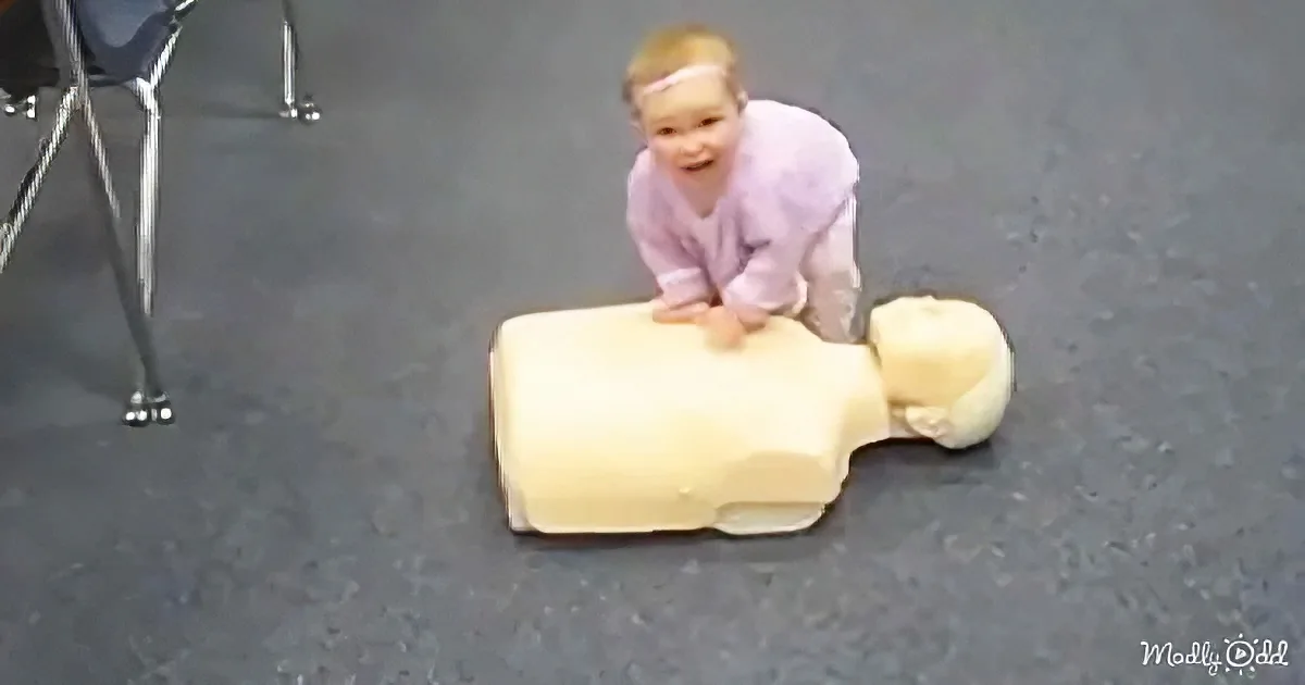 A child in a purple shirt is performing CPR on a baby mannequin on a gray floor.