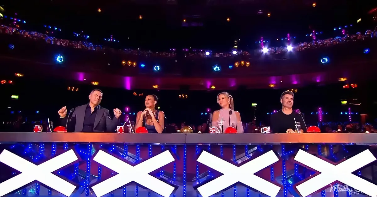 Four judges sit at a table with colorful lights and a large audience in the background.