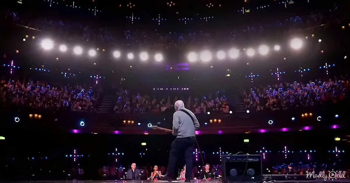 A guitarist stands on stage, facing a large, illuminated crowd in a concert setting.