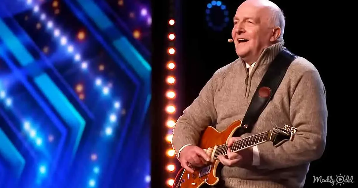 A man in a beige sweater plays a guitar on stage with colorful lights behind him.