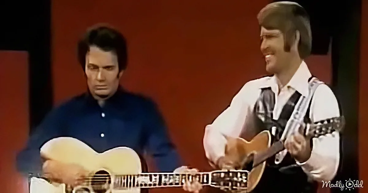 Two men play acoustic guitars on a stage with a red backdrop.