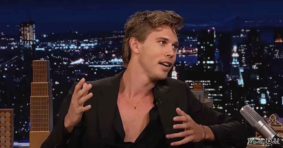 A man in a black suit gestures while speaking on a talk show set with a cityscape background.