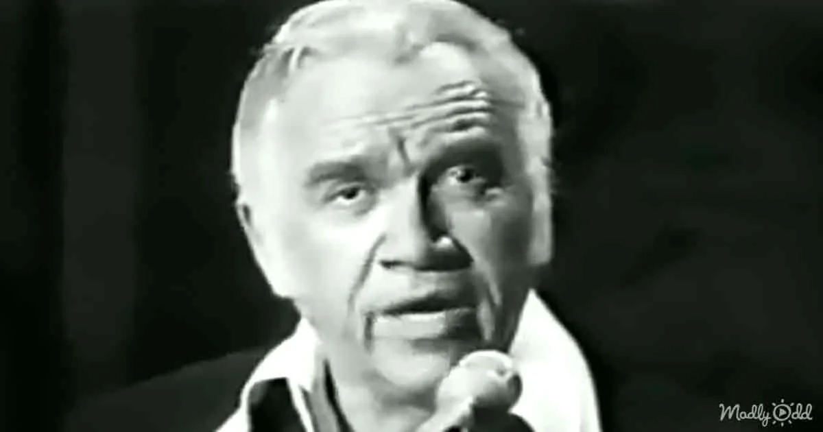 A man with a microphone, wearing a white shirt, appears to be speaking or singing in a dimly lit setting.