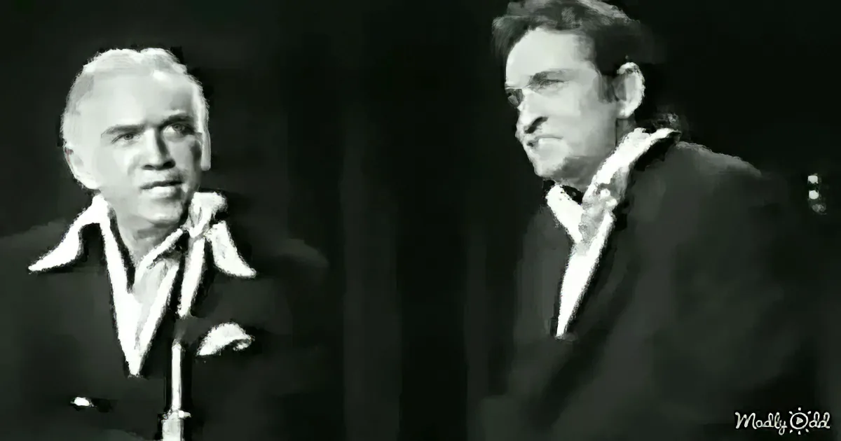 Two men in suits, one with white hair, both on stage, one smiling, one looking serious.
