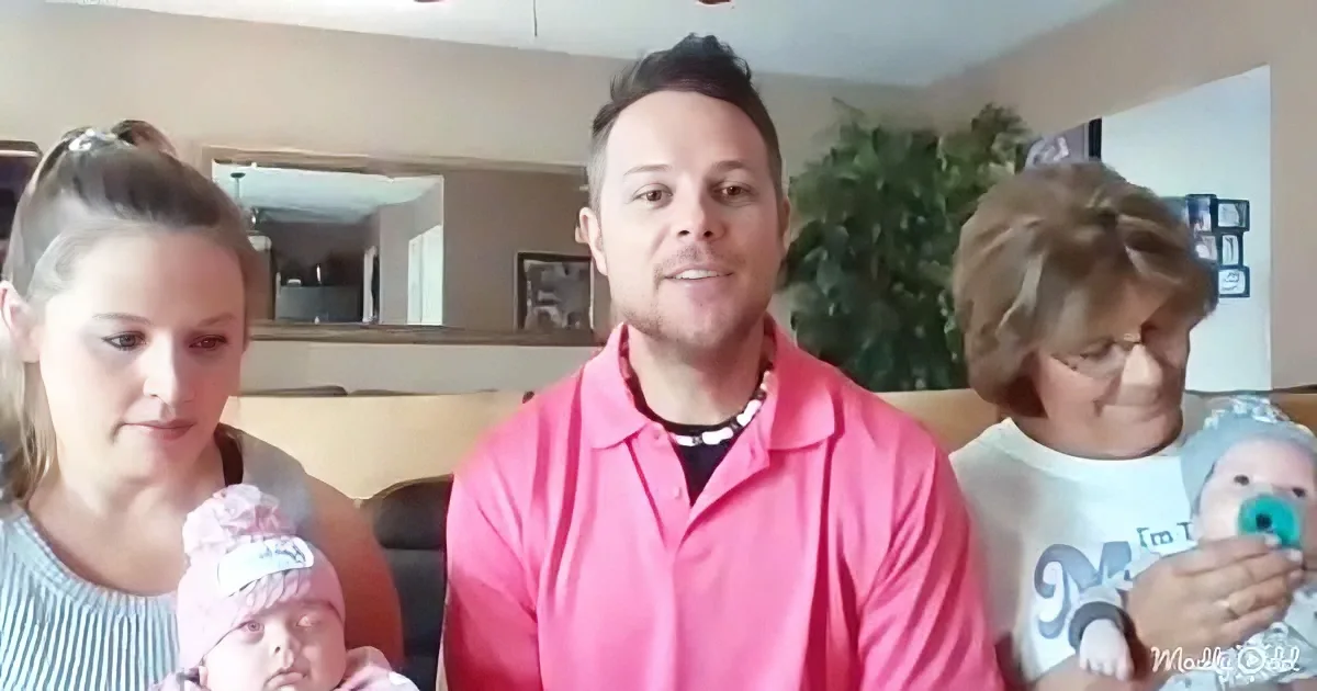 A man in a pink shirt sits between two women holding babies, in a cozy living room setting.