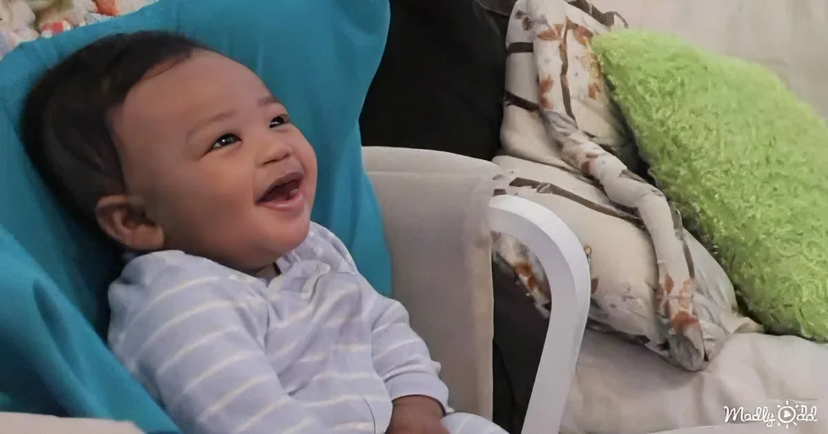 A baby in a striped onesie is laughing while sitting in a blue chair.