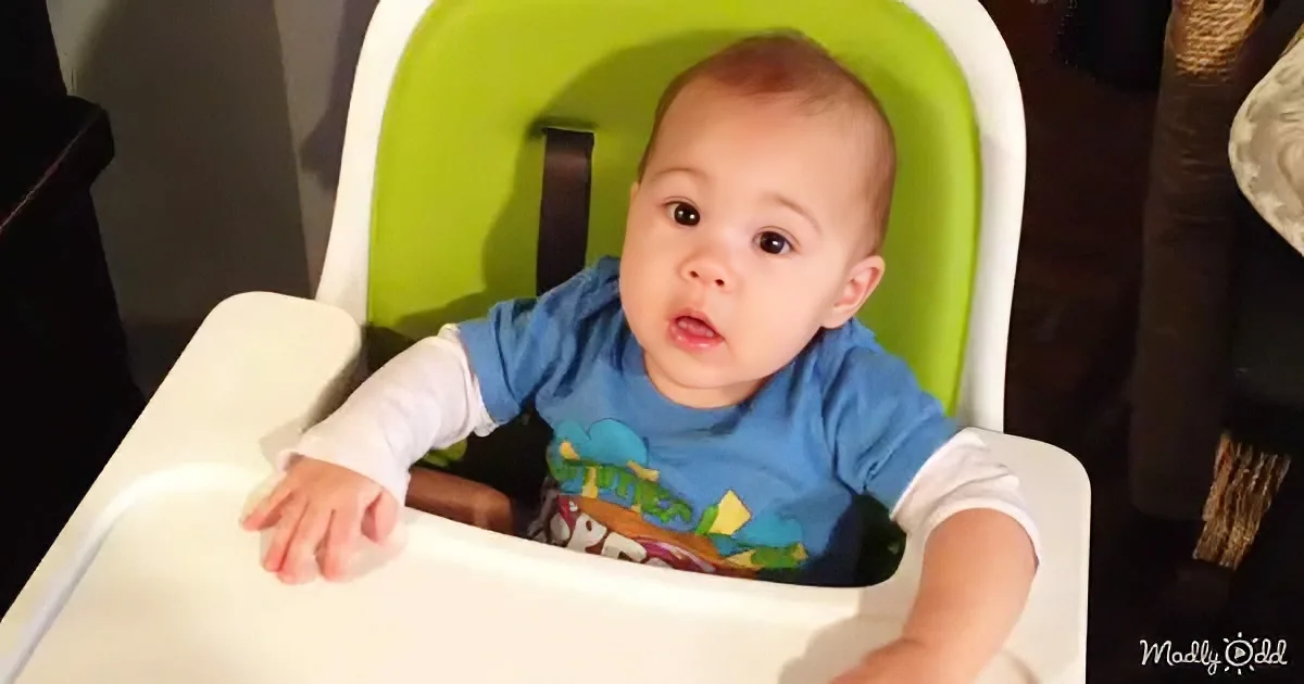 A baby in a blue shirt sits in a green and white high chair, looking up with a curious expression.
