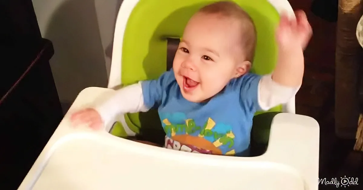 A baby in a high chair smiles widely, wearing a blue shirt with a colorful design.