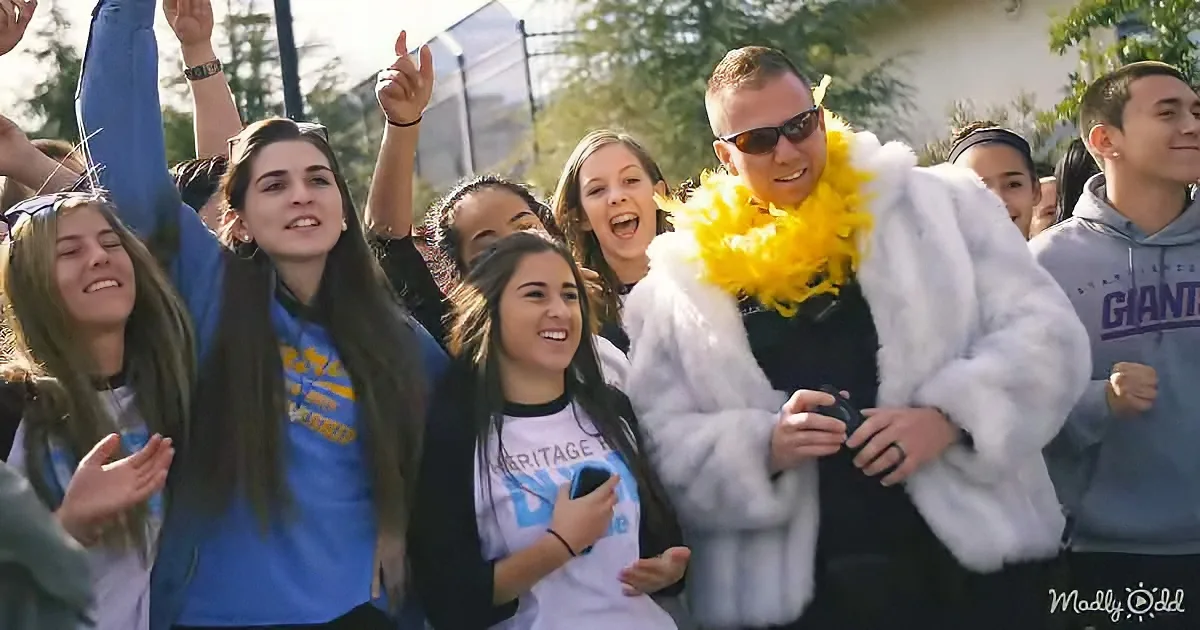 A man in a fur coat and yellow boa stands with a group of excited students, likely at a school event.