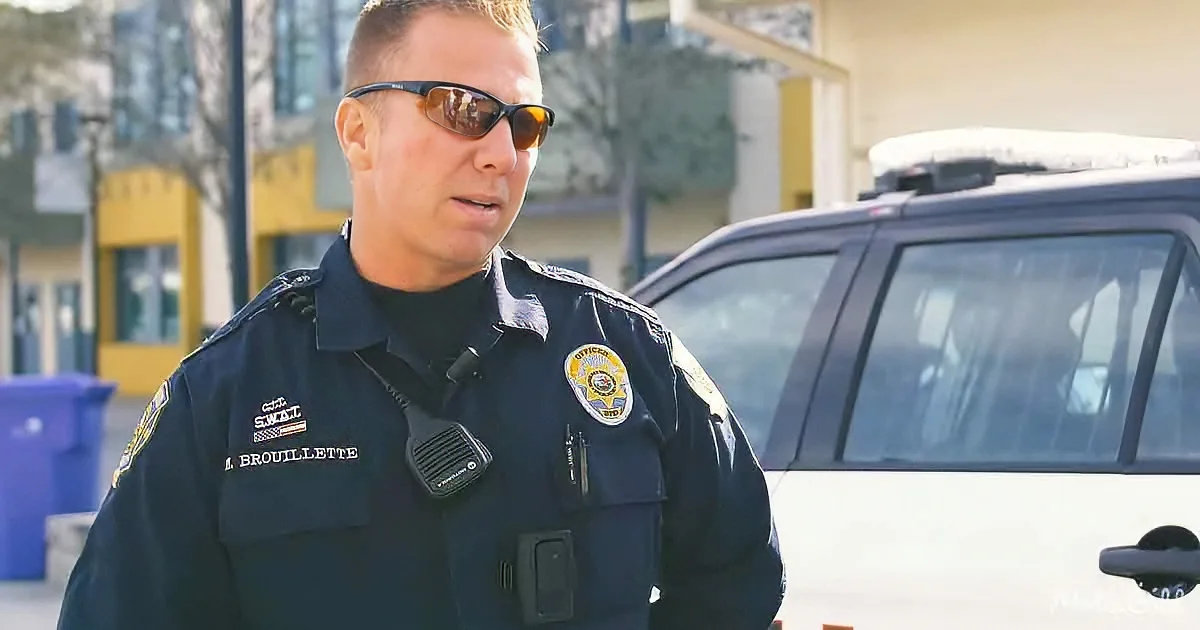 A police officer in uniform stands in front of a police car, wearing sunglasses and a badge.