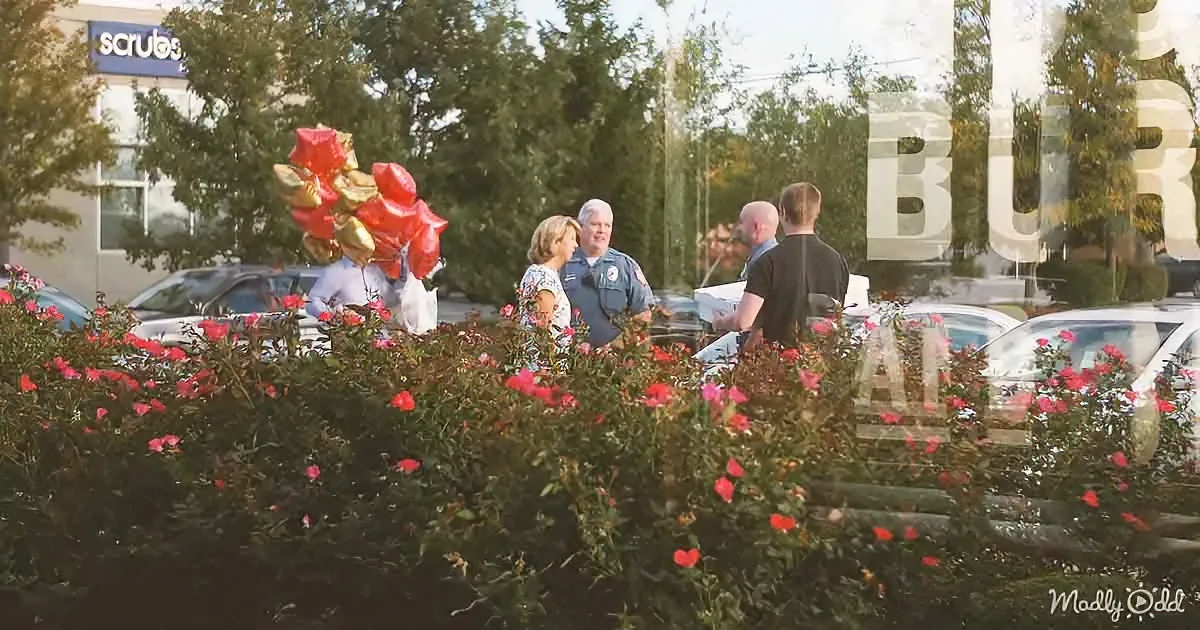 A group of people, including a police officer, stand near a bush with red flowers and balloons, in a parking lot.