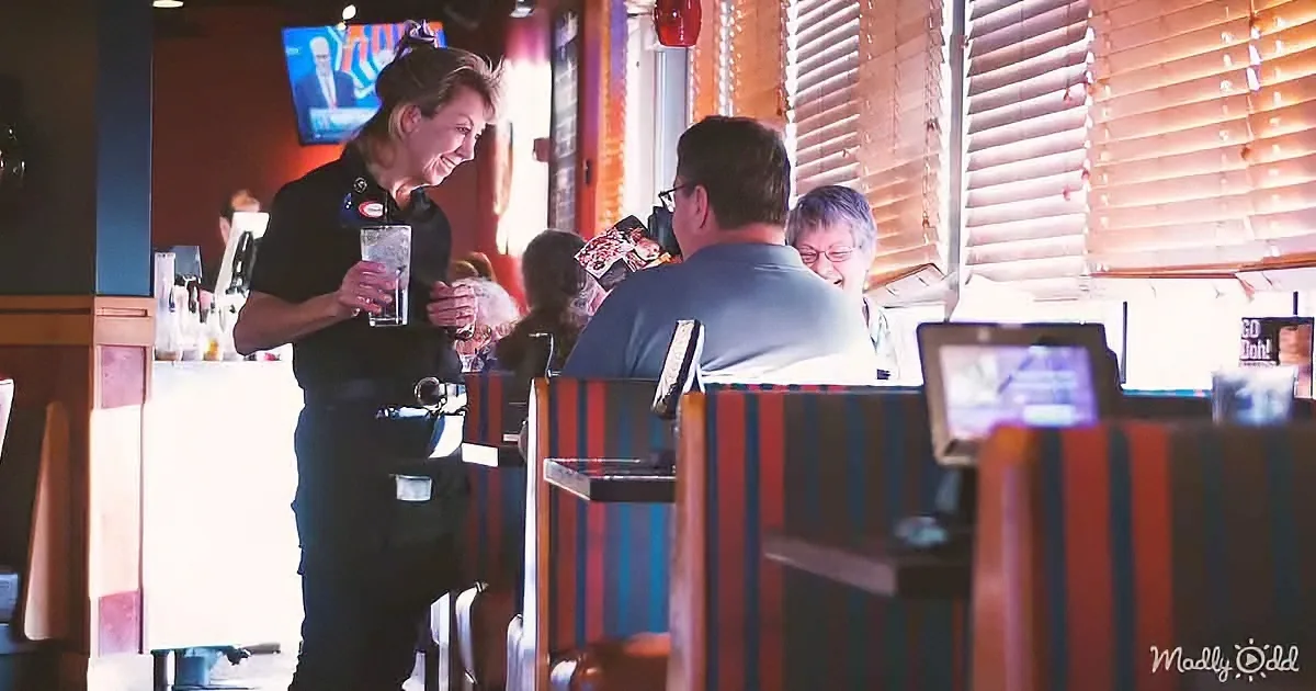 A waitress in a black uniform serves a drink to a man at a restaurant with striped booth seats.