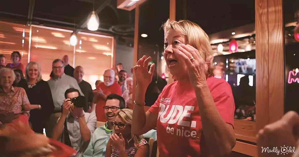 A woman in a red "Dude, be nice" shirt claps enthusiastically in a lively crowd.