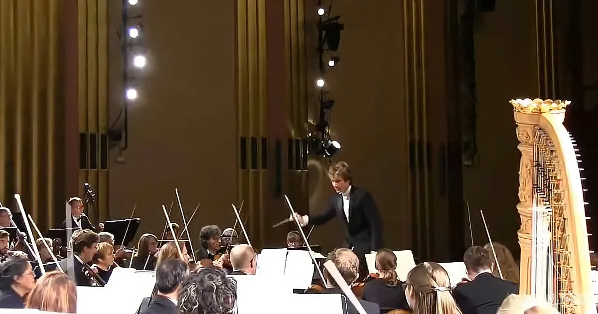 Conductor leads orchestra with baton in formal attire, surrounded by musicians and harp.
