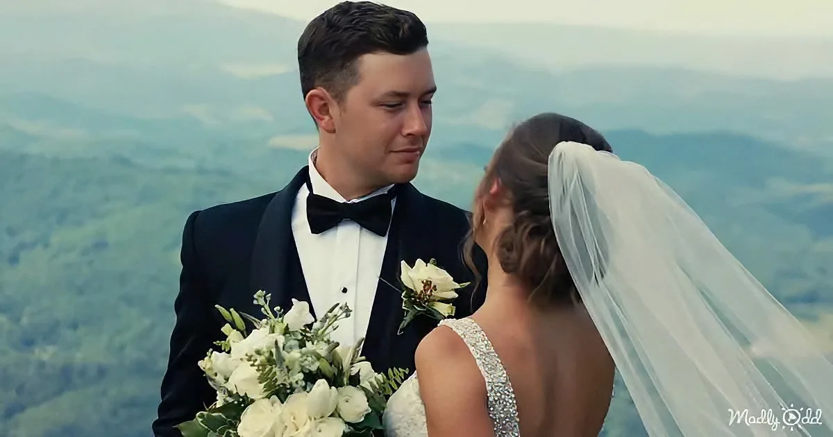 A bride and groom stand together, the bride in a white dress and veil, the groom in a tuxedo, both holding bouquets...