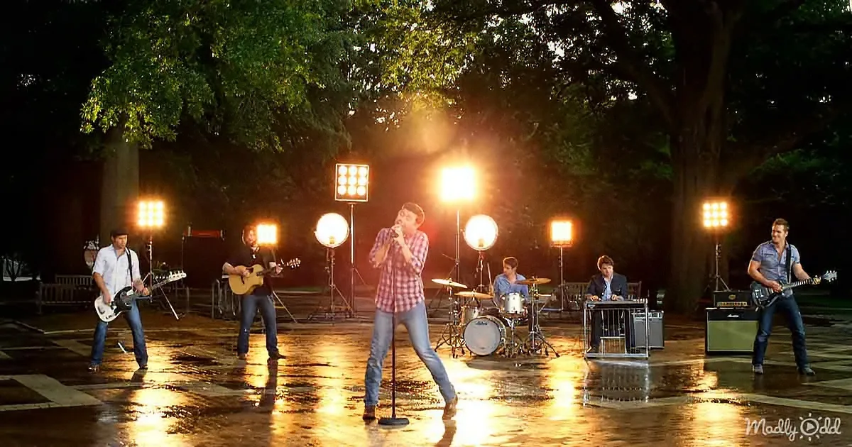 A man in a plaid shirt sings into a microphone on a wet stage with a band and bright lights.