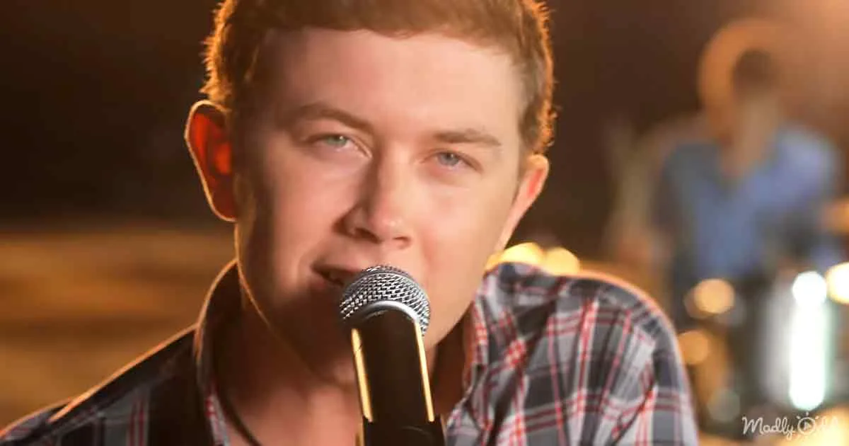 A person with short red hair sings into a microphone, wearing a plaid shirt, in a dimly lit setting.