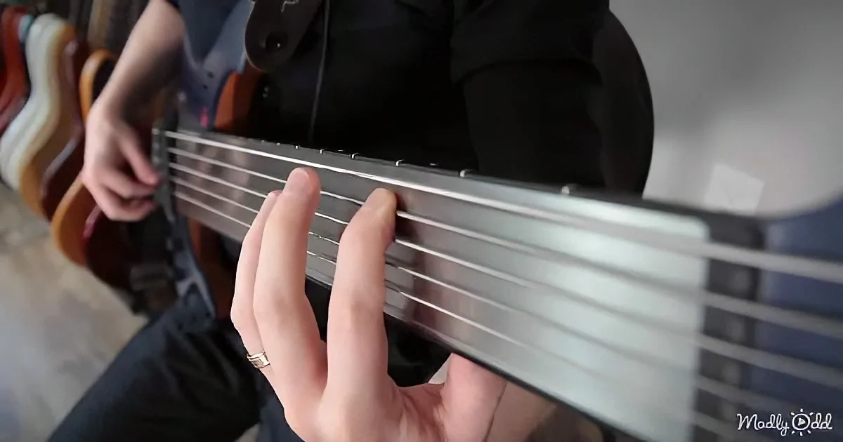 A person plays a six-string bass guitar, focusing on the fretboard with their left hand.