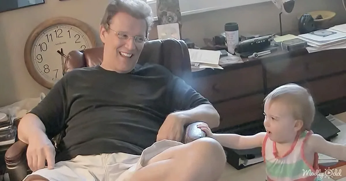 A man and a child are sitting together, smiling, in a cozy room with a clock and desk in the background.