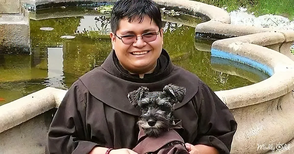 A person in a brown robe holds a small black dog, smiling near a stone fountain with green water.