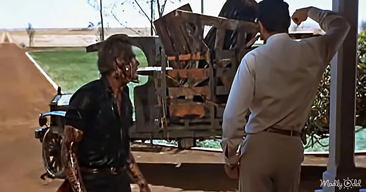 A man in a black leather jacket stands facing another man in a white shirt, with a motorcycle and a wooden cart in the...