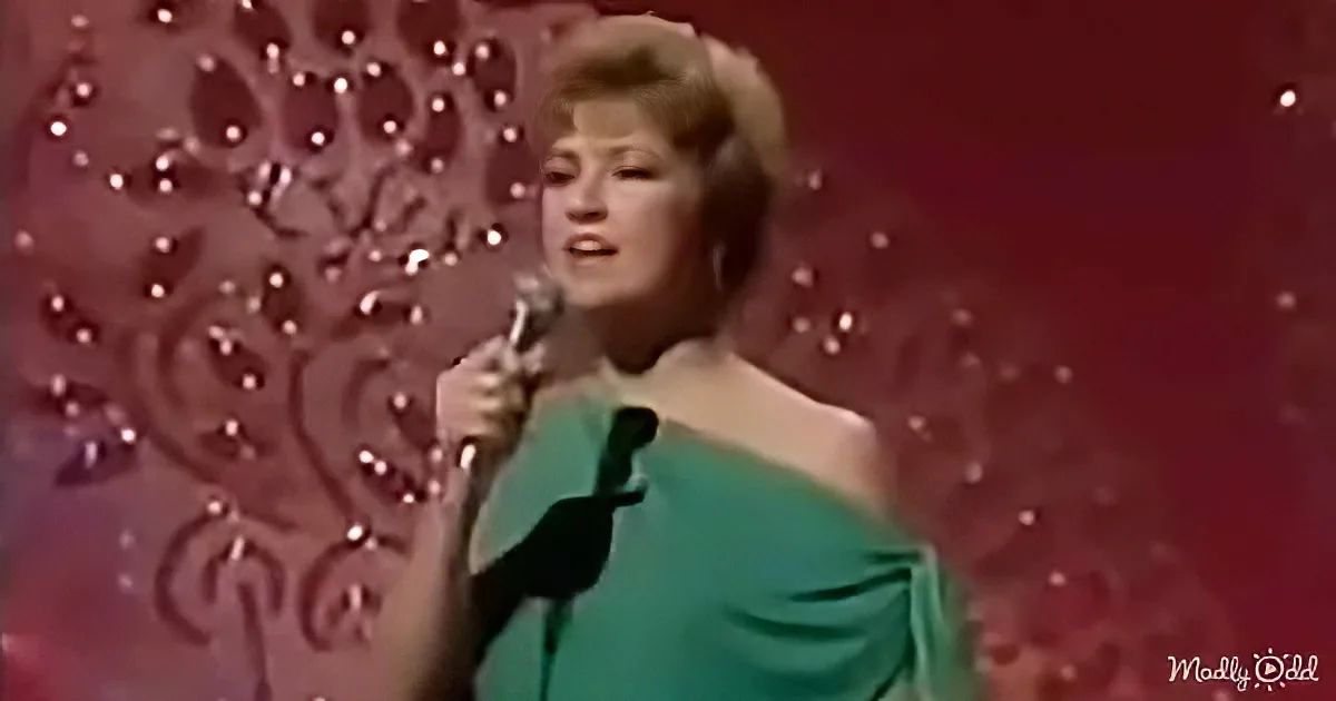 A woman in a green dress sings into a microphone on stage with a red, patterned backdrop.