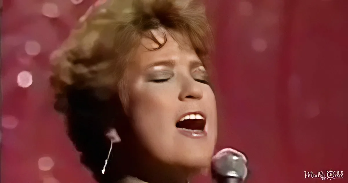 A woman with short, curly hair sings into a microphone against a red, sparkly background.