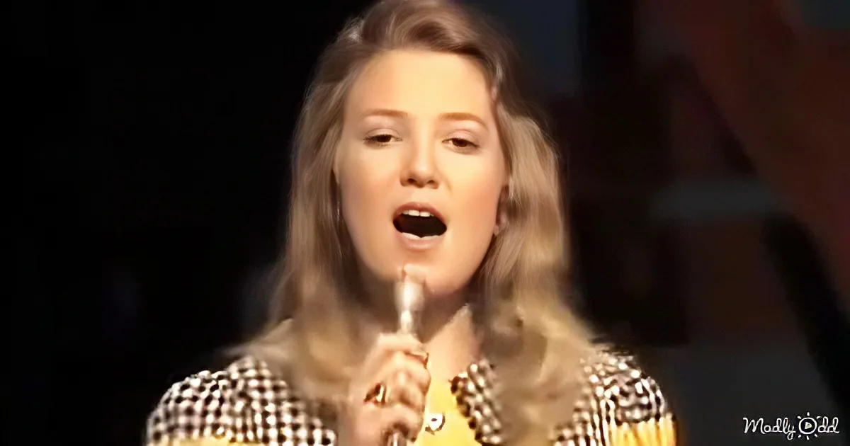 A person with long, wavy hair sings into a microphone, wearing a checkered top.