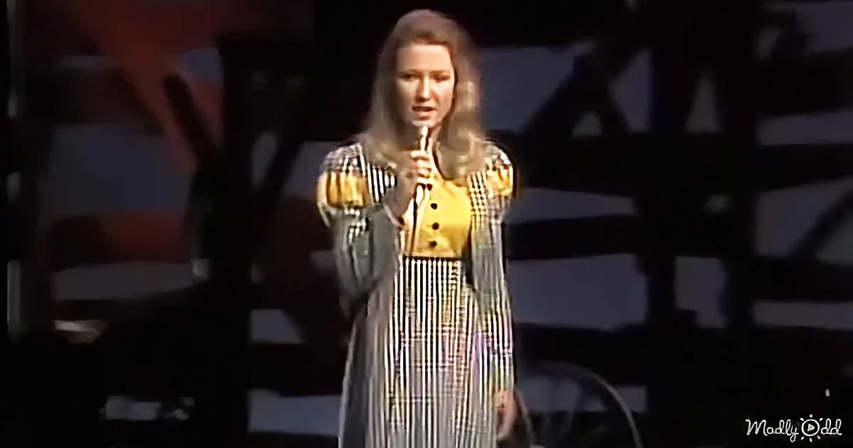 A woman in a striped dress holds a microphone on stage, illuminated by a spotlight.