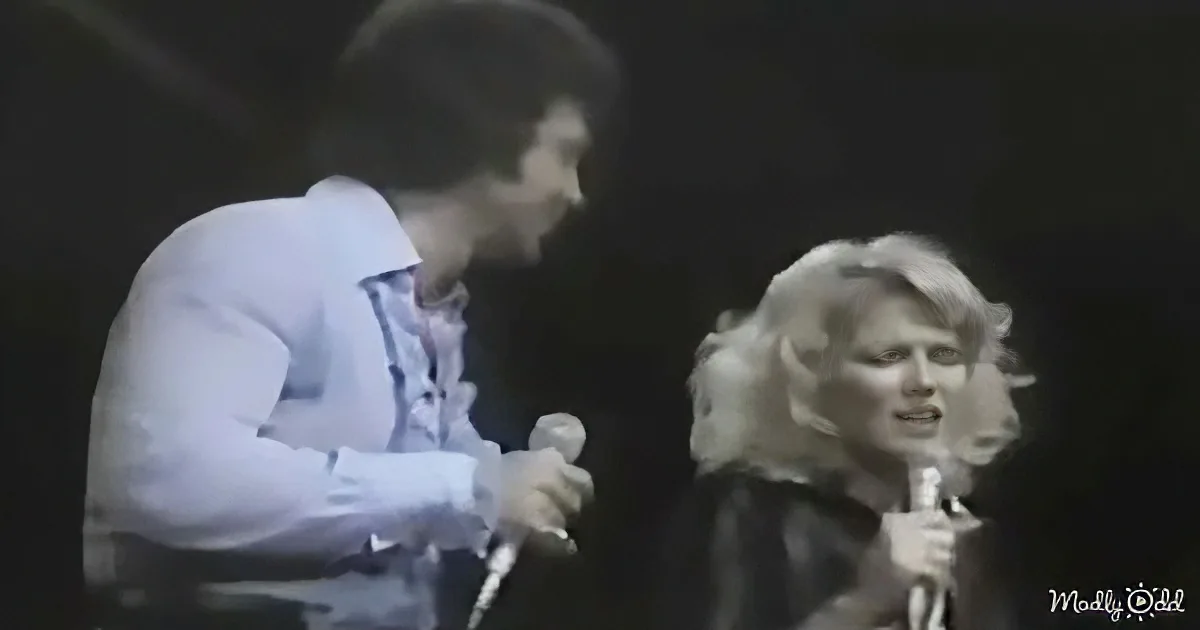 A man and woman are singing on stage, both holding microphones, with the man in a light shirt and the woman in a dark outfit.