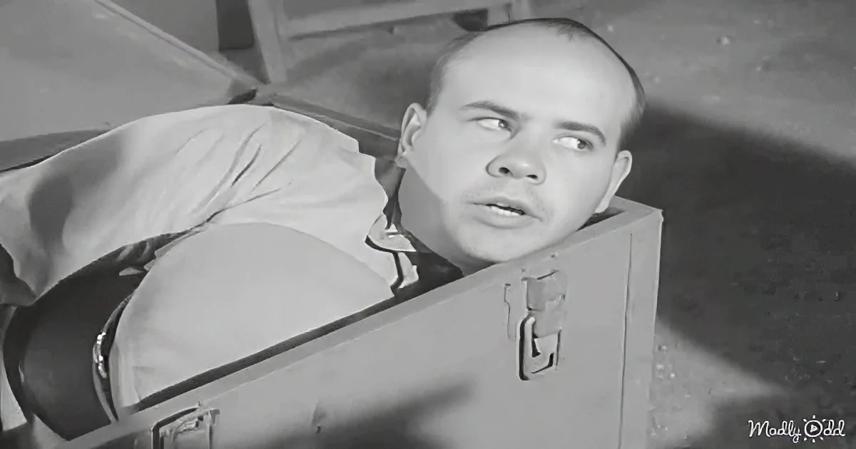 A man in a suit lies on a suitcase, looking up with a concerned expression.