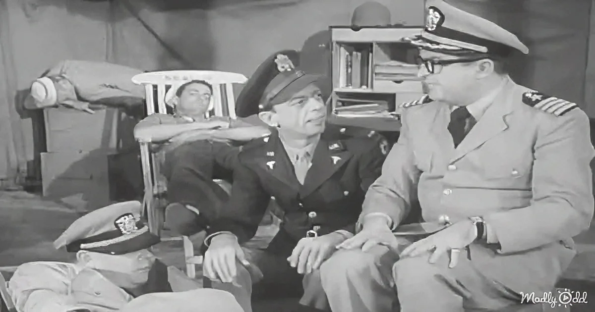 Two men in military uniforms are seated, engaged in conversation, with another man lying in a chair in the background.