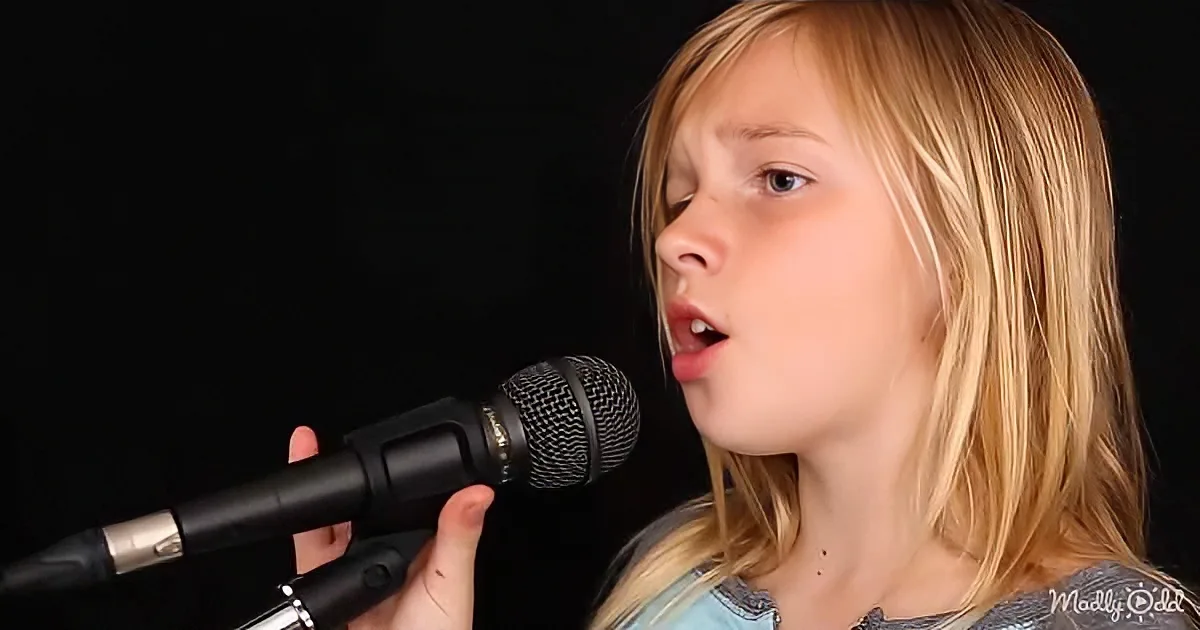 A young girl with blonde hair sings into a microphone against a dark background.