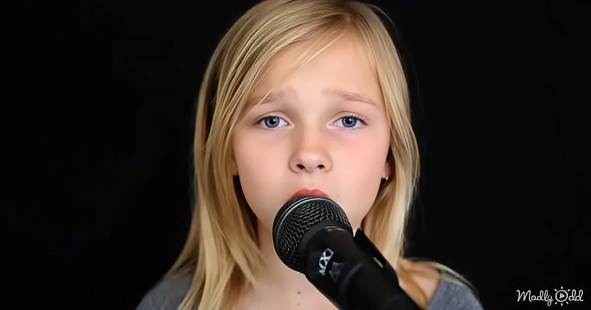 A young girl with blonde hair sings into a microphone against a dark background.