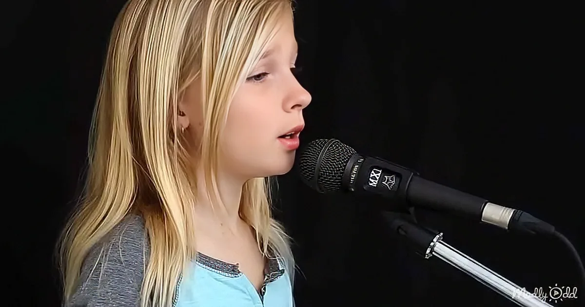 A young person with long blonde hair sings into a black microphone against a dark background.